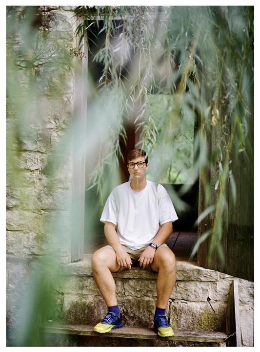 A man wearing glasses, a white t-shirt, and shorts sits on stone steps, framed by hanging willow branches.
