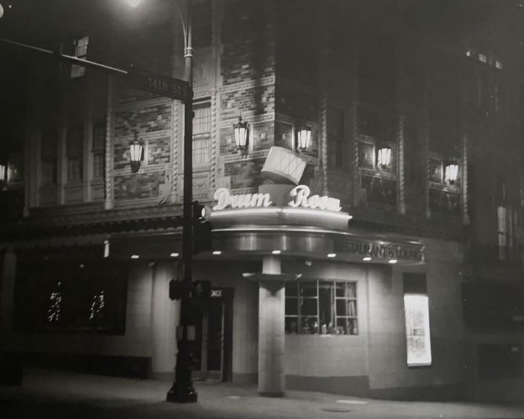 A street corner building with a neon sign for "Drum Room Restaurant & Lounge" is illuminated at night.