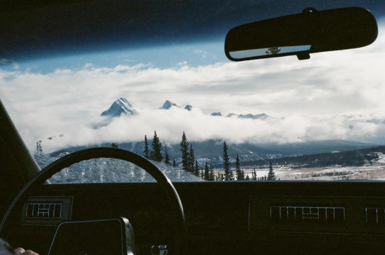 A mountain range is visible through a car windshield, with clouds obscuring the lower peaks and a steering wheel in the foreground.