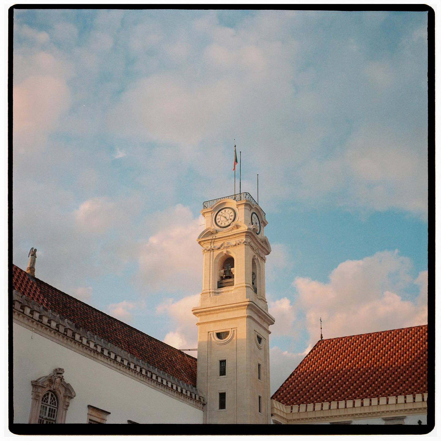 A tall clock tower with a flag on top stands against a cloudy sky, flanked by red-tiled rooftops.