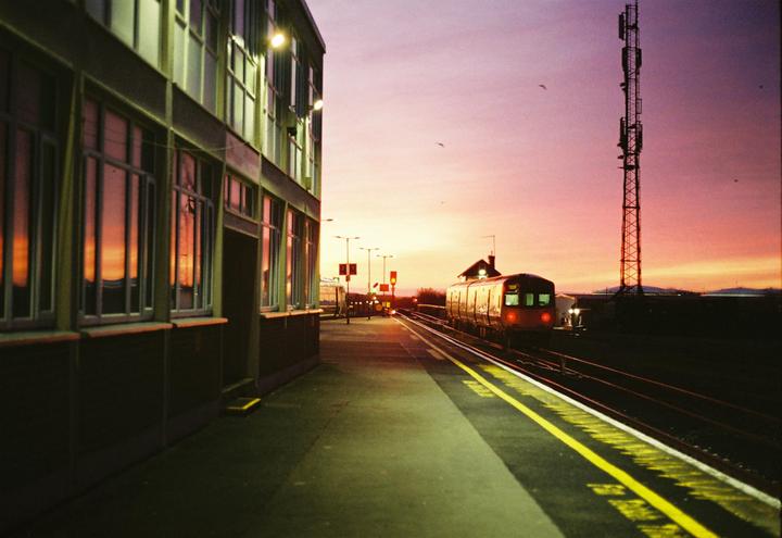 A train is stopped at a station platform at sunset, with a building on the left and a large antenna tower on the right.