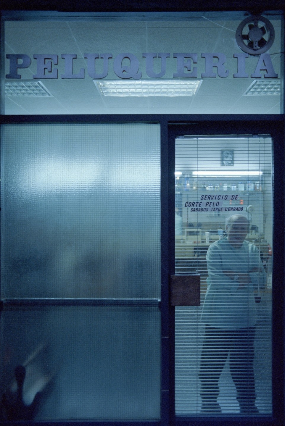 A man stands inside a barber shop with a sign that reads "Peluqueria" above the window.