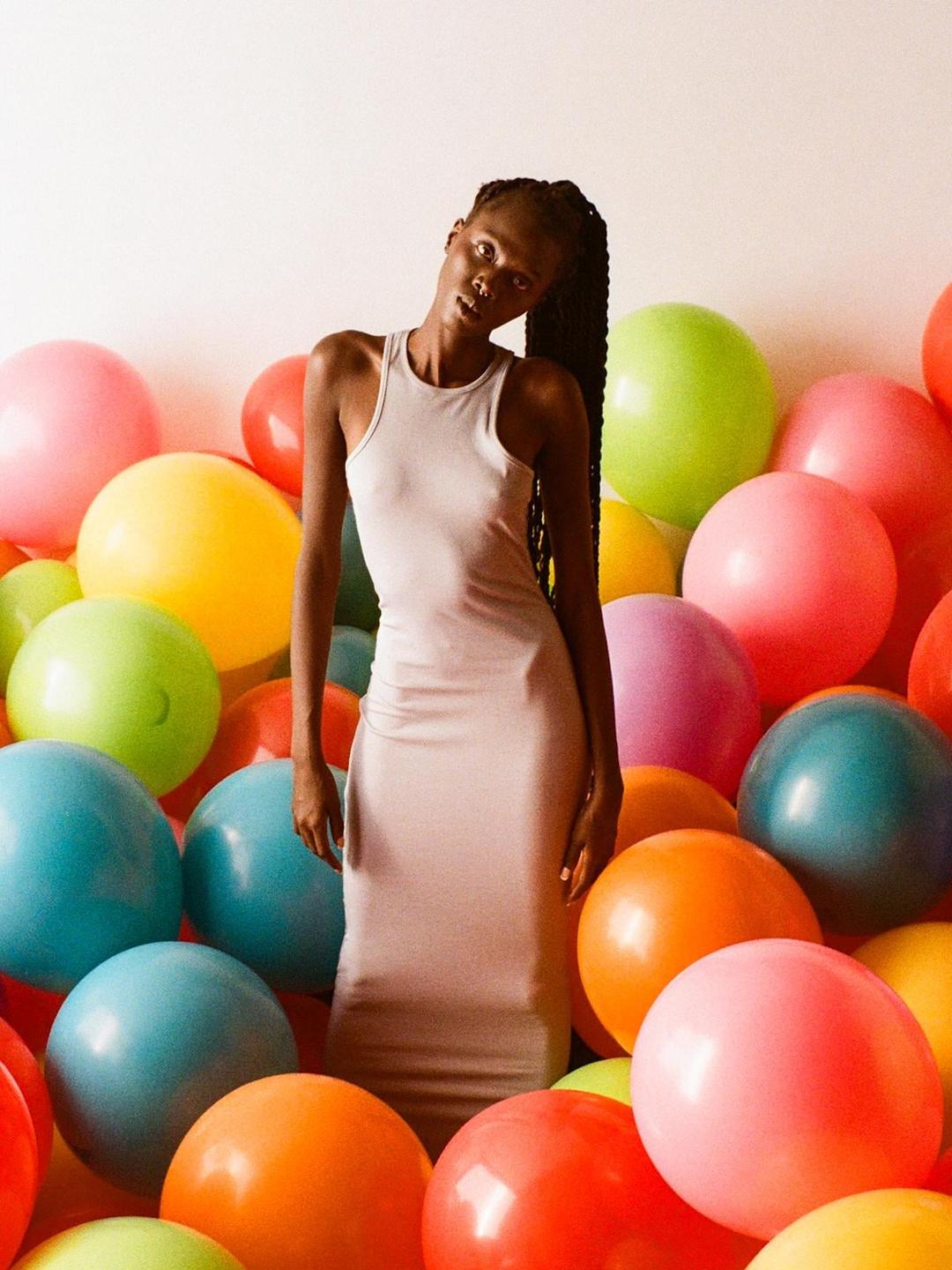 A woman in a light-colored dress stands among many colorful balloons.