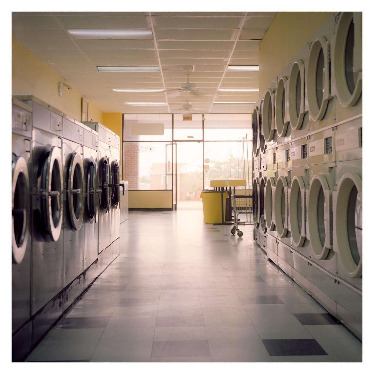 A long row of washing machines lines both sides of a laundromat, leading to an open door at the far end.