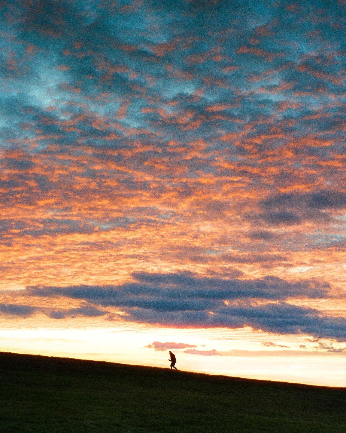 A person walks up a grassy hill under a dramatic sunset sky with orange and blue clouds.