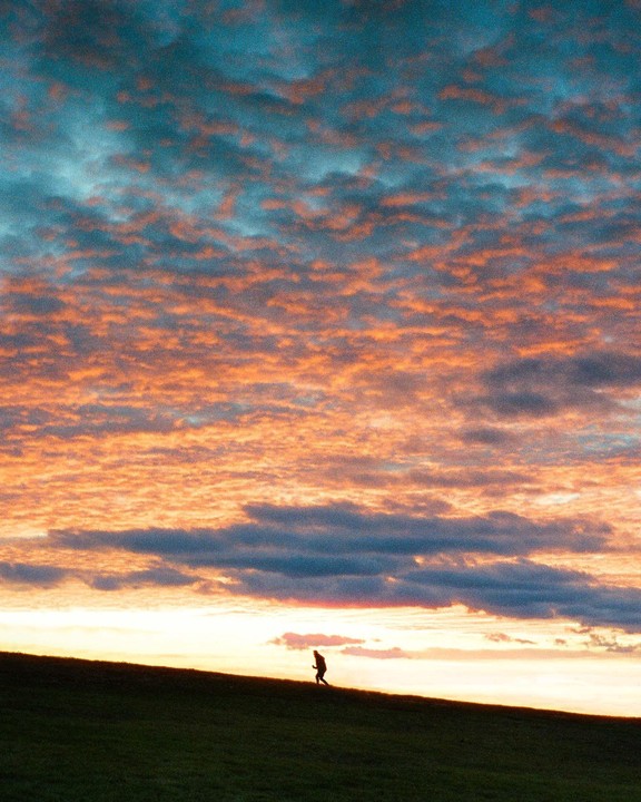 A person walks up a grassy hill under a dramatic sunset sky with orange and blue clouds.