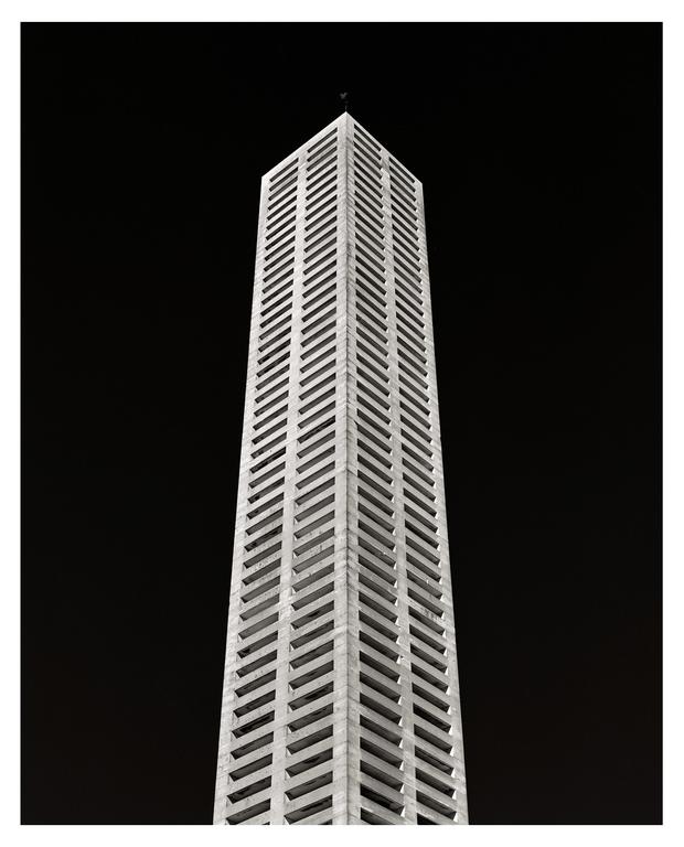 A tall, narrow building with a repeating pattern of windows rises against a dark sky.