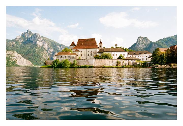 A large building with a red roof sits on the edge of a lake, with mountains in the background.