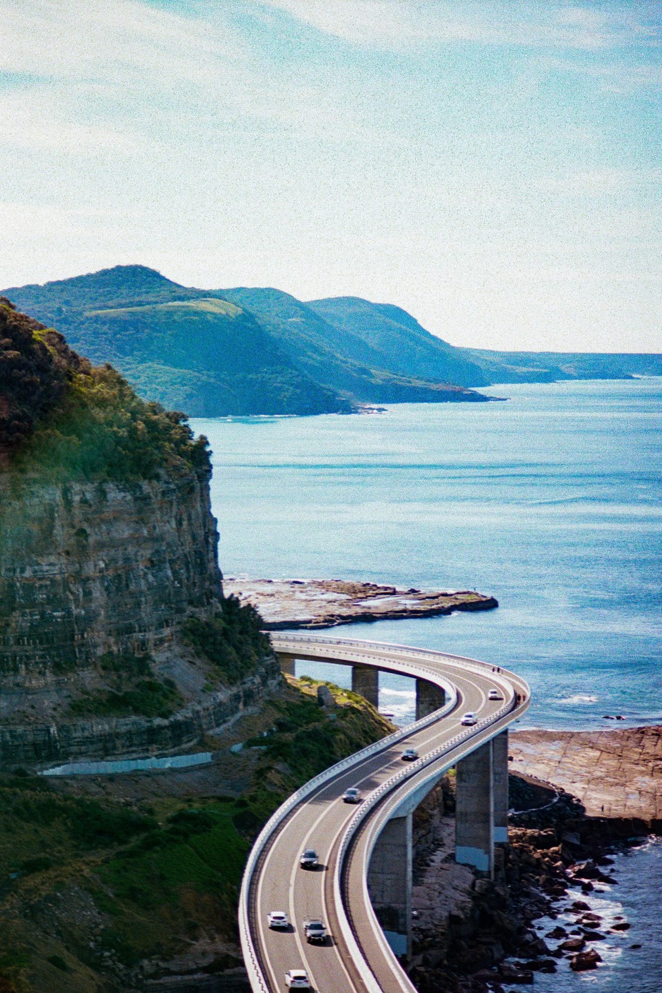 A winding coastal road with cars on it is built into a cliff face overlooking the ocean, with mountains in the distance.