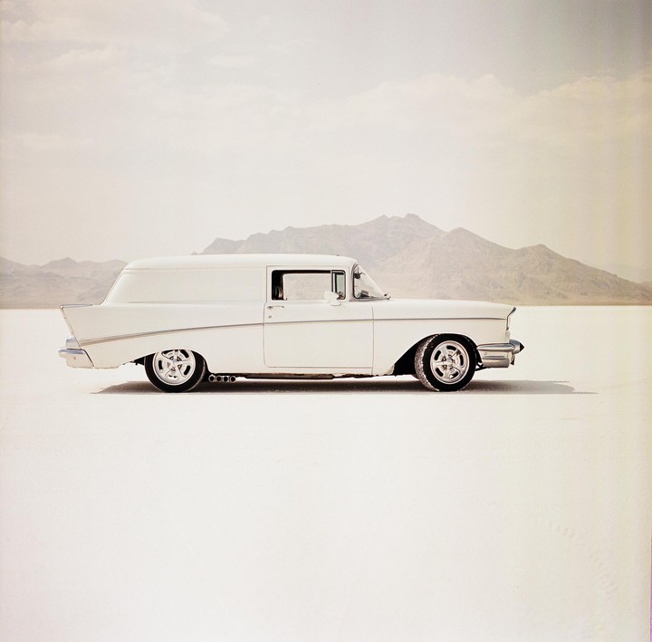 A white vintage car is parked on a vast, flat, white expanse with mountains in the background.