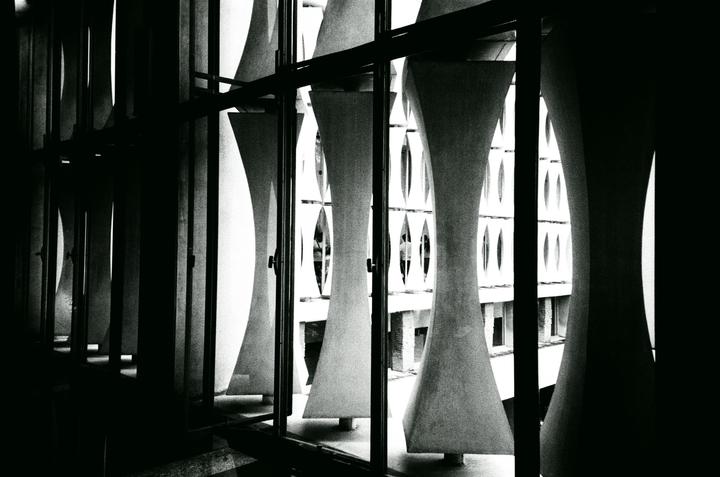 A dark interior looks out through a window with vertical concrete fins at a building with oval windows.