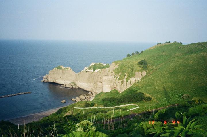A winding road leads down a grassy hillside to a rocky beach and the ocean, with a large rock formation jutting out into the water.