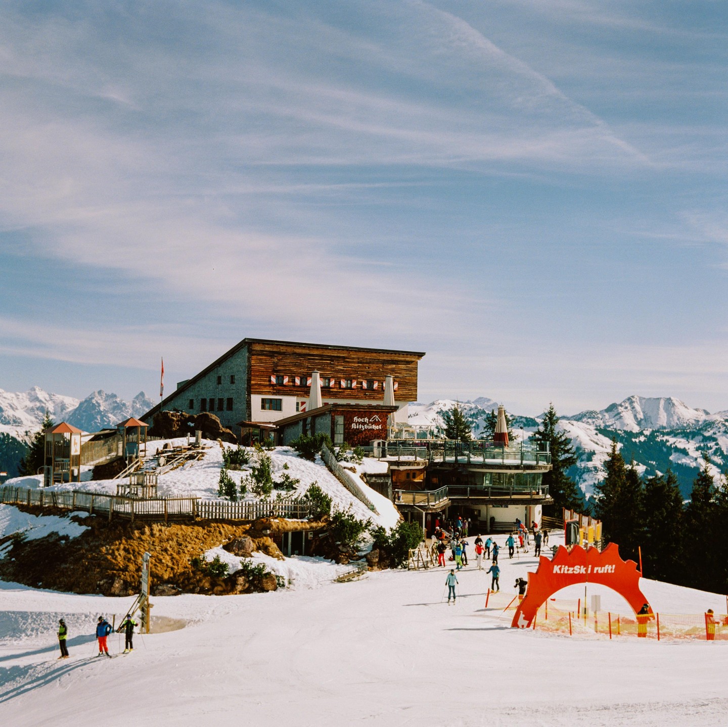 A ski resort building with a large wooden facade sits on a snowy mountain with skiers on the slope in the foreground.