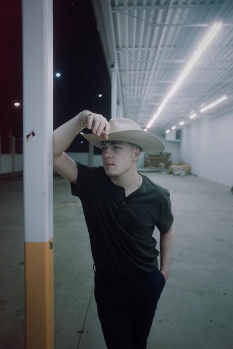 A man in a cowboy hat leans against a pole in a parking garage at night.