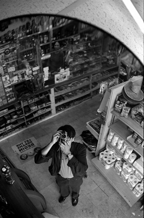 A man takes a self-portrait in a store mirror, with a woman visible in the reflection behind him.