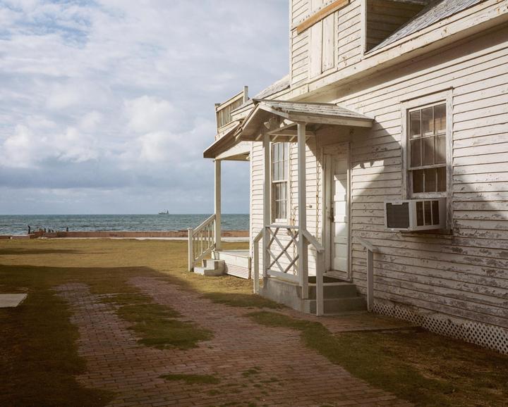A weathered white house with a porch and an air conditioner in a window stands beside a brick path and a grassy area, with the ocean and a distant ship visible under a cloudy sky.