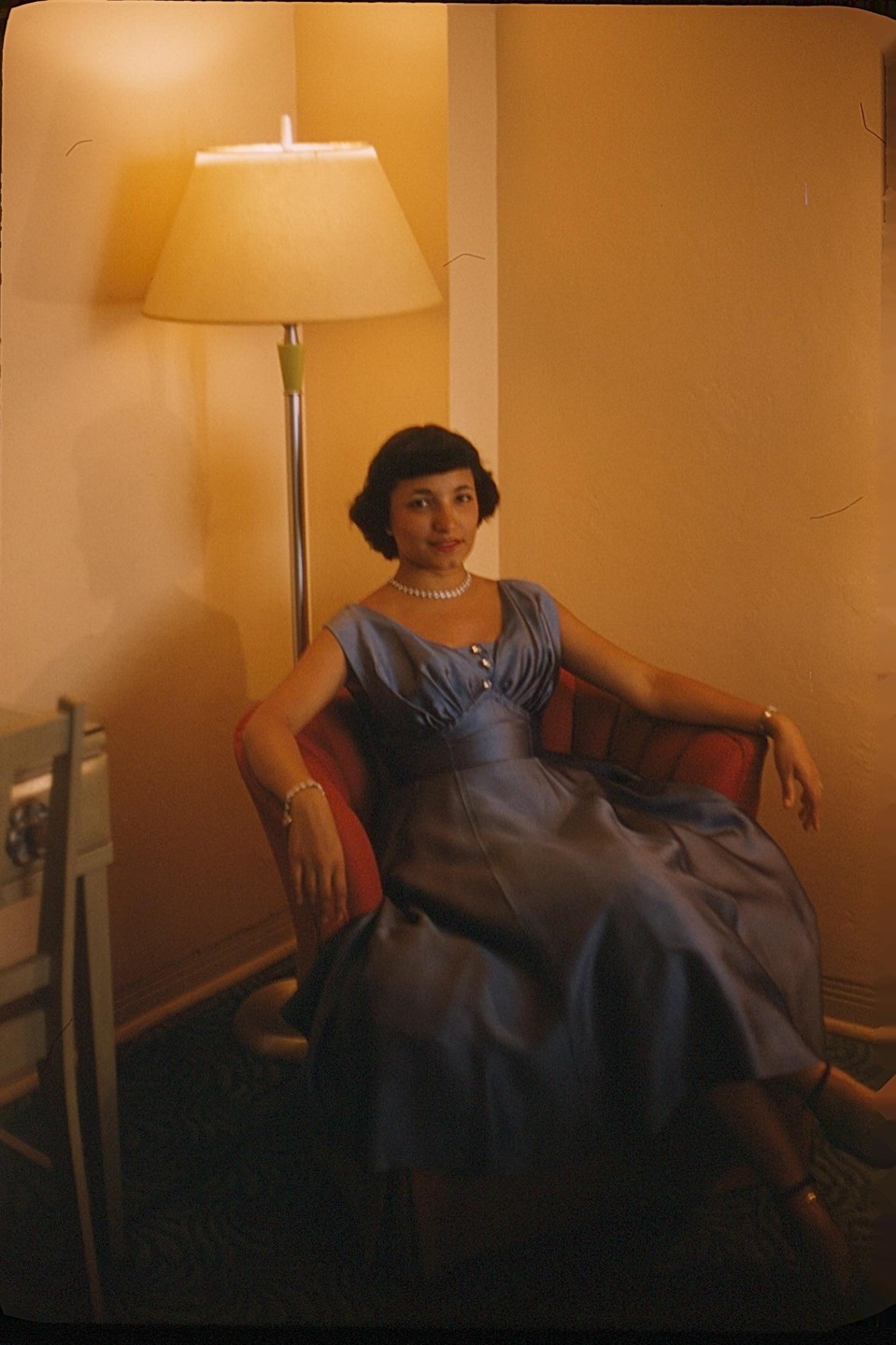 A woman in a blue dress and pearl necklace sits in a red armchair next to a floor lamp.
