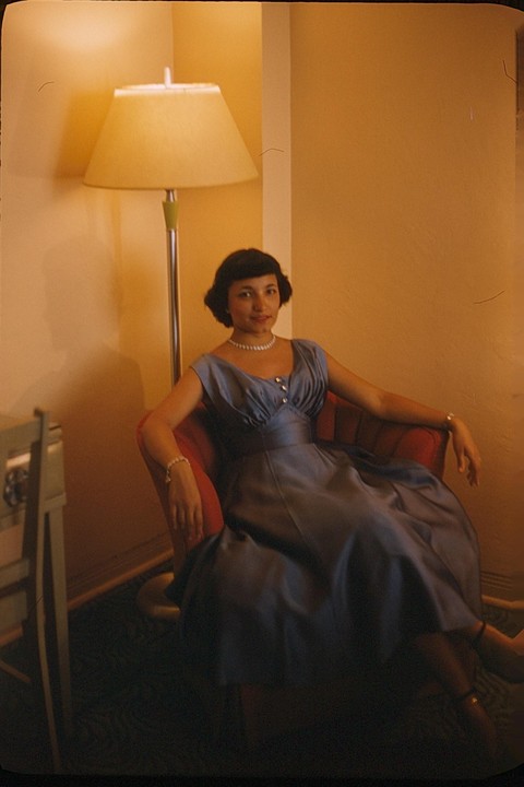 A woman in a blue dress and pearl necklace sits in a red armchair next to a floor lamp.