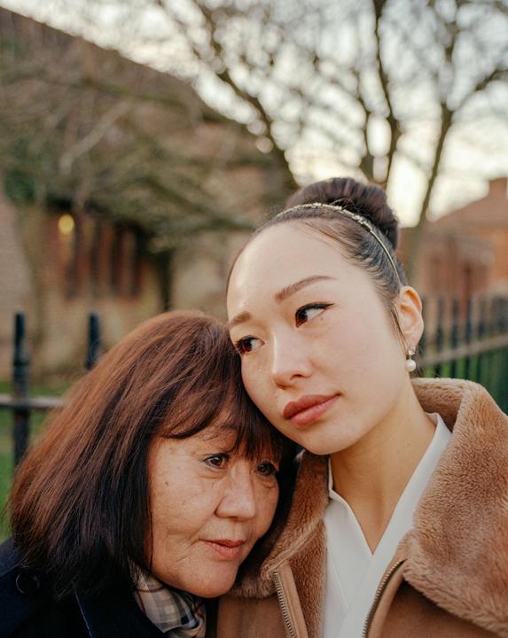 A woman rests her head on an older woman's shoulder, both looking in the same direction.