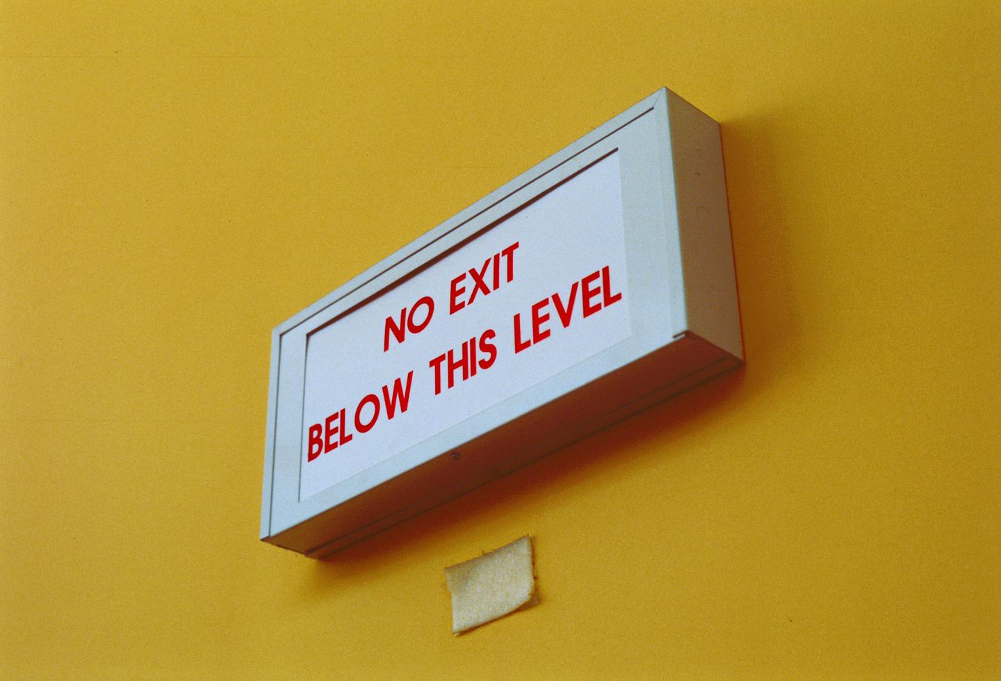 A white sign with red text reading "NO EXIT BELOW THIS LEVEL" is mounted on a bright yellow wall.