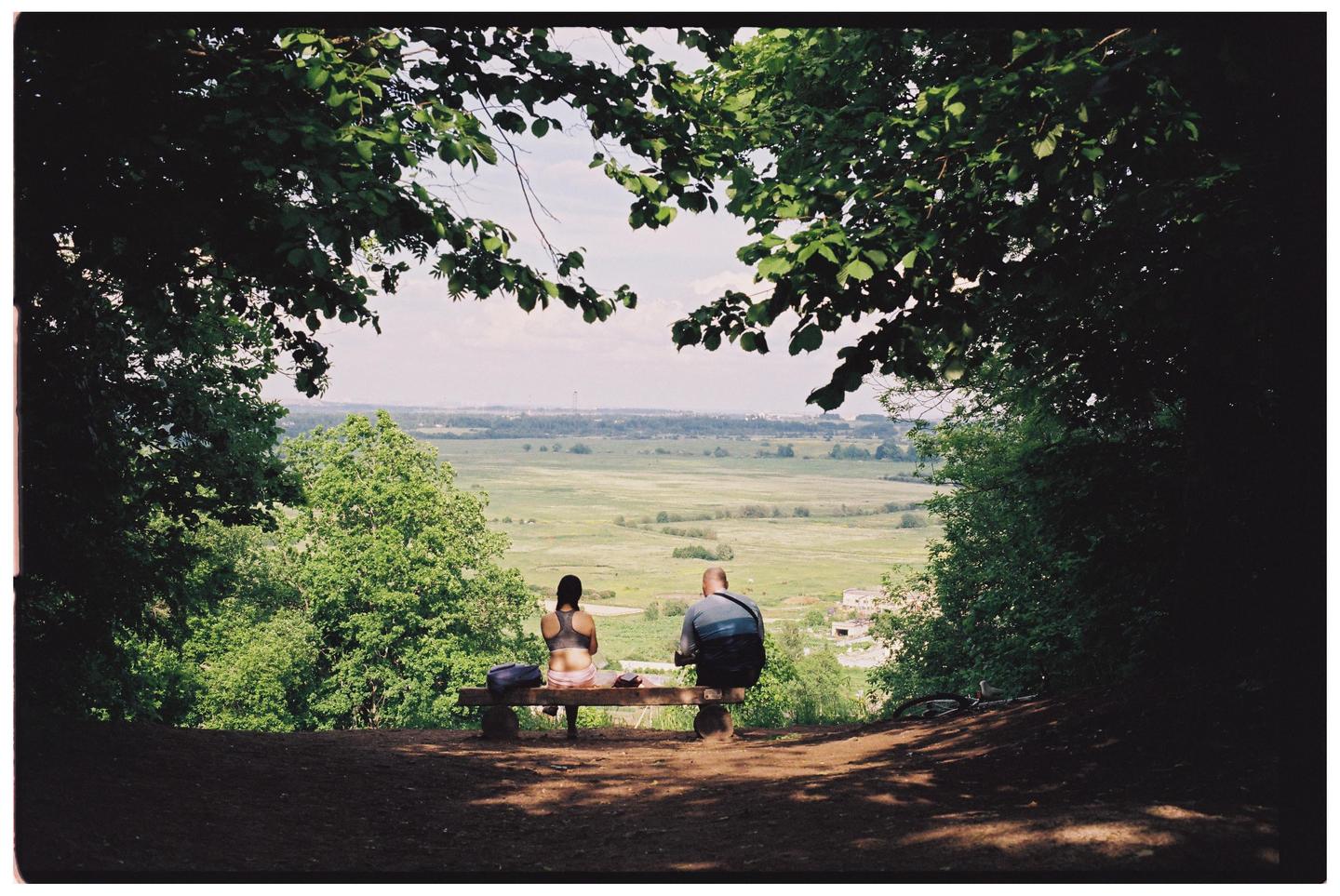 A couple sits on a bench overlooking a vast green landscape under a bright sky, framed by dark foliage.