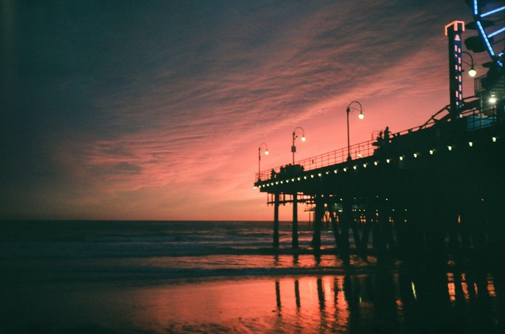 A pier with lights is silhouetted against a vibrant pink and orange sunset over the ocean.
