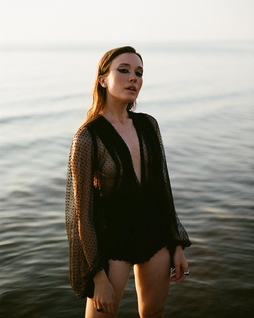 A woman stands in the sea wearing a sheer black top and black bottoms.