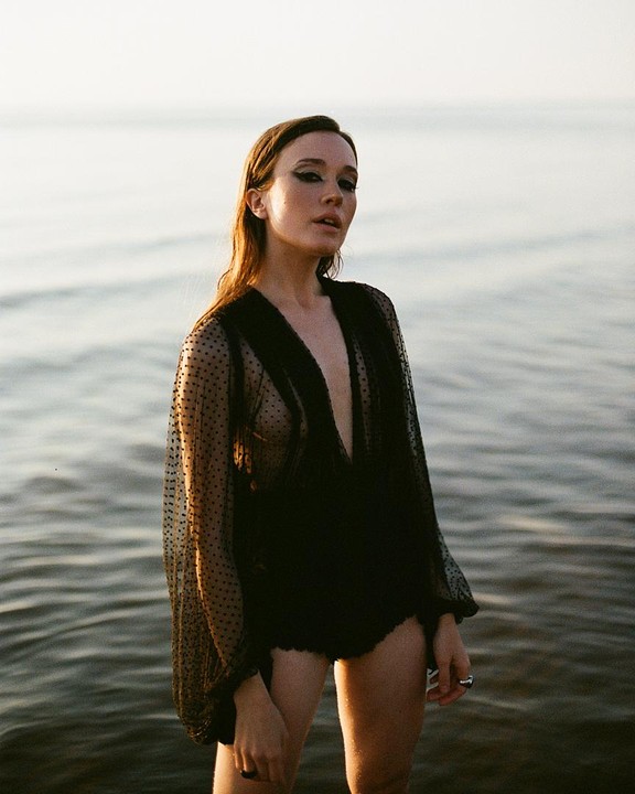 A woman stands in the sea wearing a sheer black top and black bottoms.