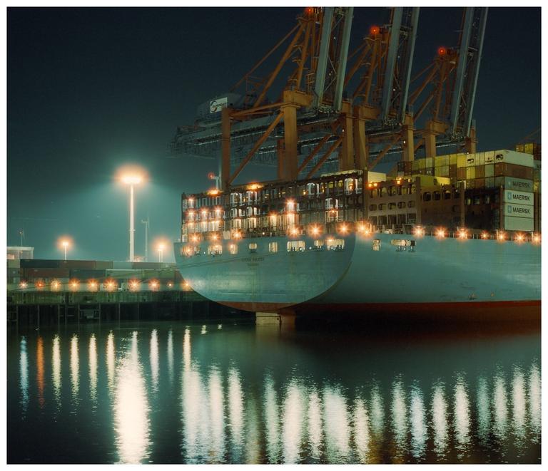 Two large cargo ships are docked at a port at night, with bright lights reflecting on the water.