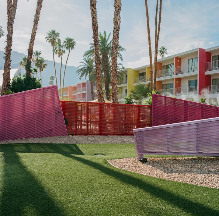 A colorful hotel building stands behind a vibrant pink and red fence, with palm trees and mountains in the background.