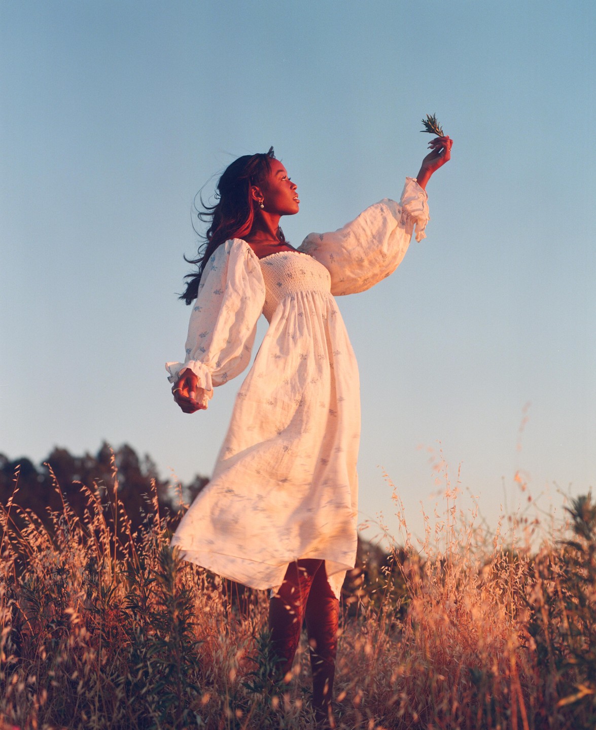 A woman in a white dress stands in a field of tall grass, holding a sprig of leaves towards the sky.