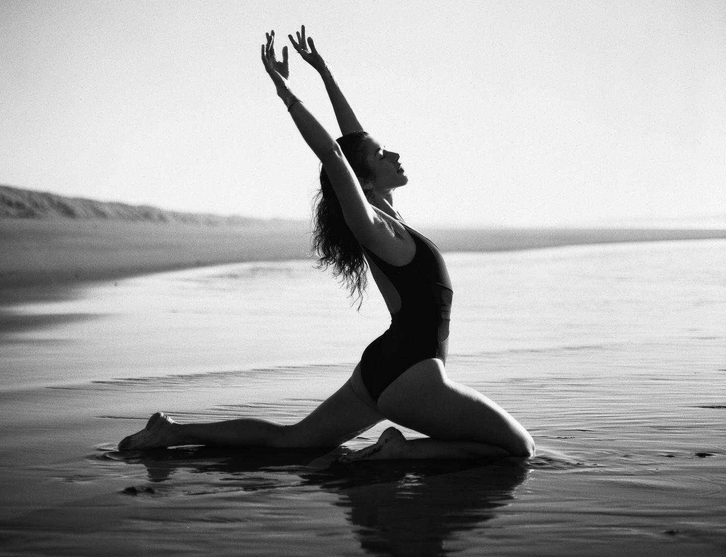 A woman in a swimsuit kneels in shallow water on a beach with her arms raised.