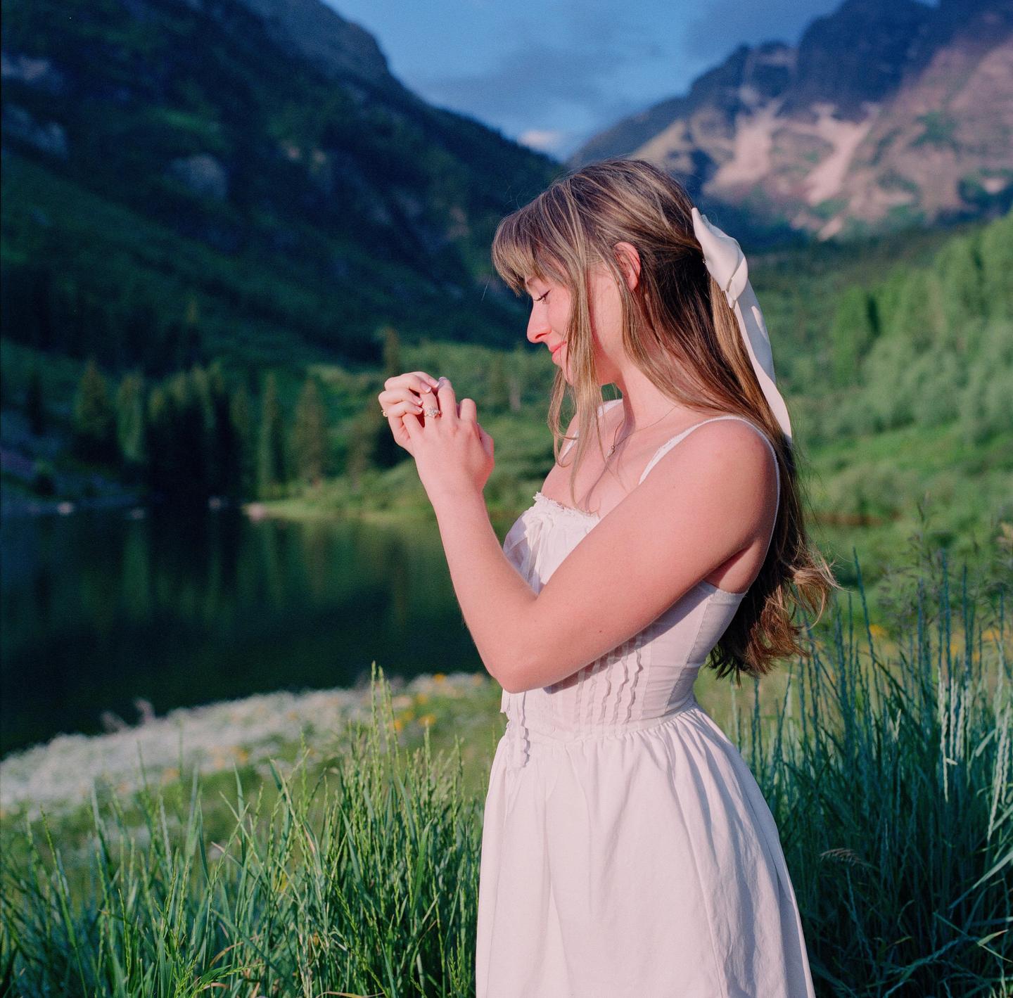 A woman in a white dress stands in a grassy field with mountains and a lake in the background.