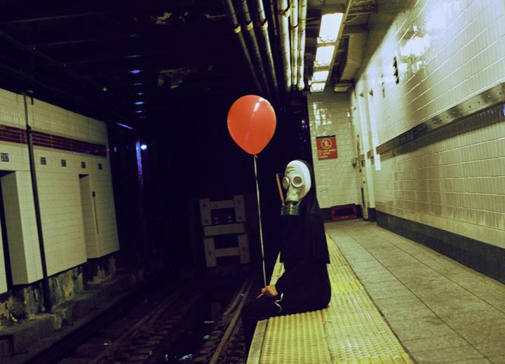 A person wearing a gas mask and a dark robe sits on a subway platform holding a red balloon.