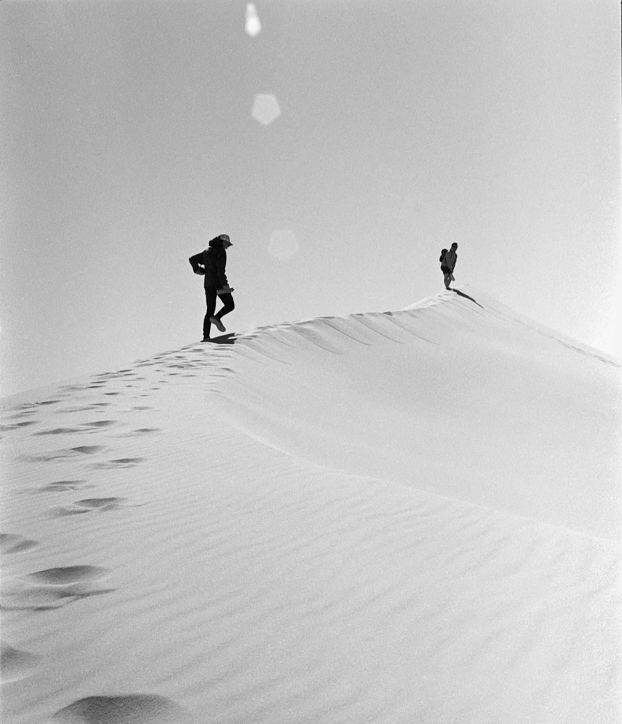 Two people walk along the ridge of a sand dune, leaving footprints in the sand.