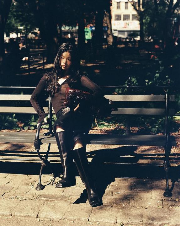 A woman sits on a park bench in dappled sunlight, holding a red bag.