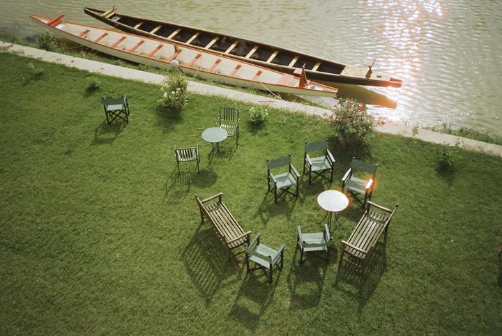 Two long boats are docked on the bank of a river next to a grassy area with several chairs and tables.