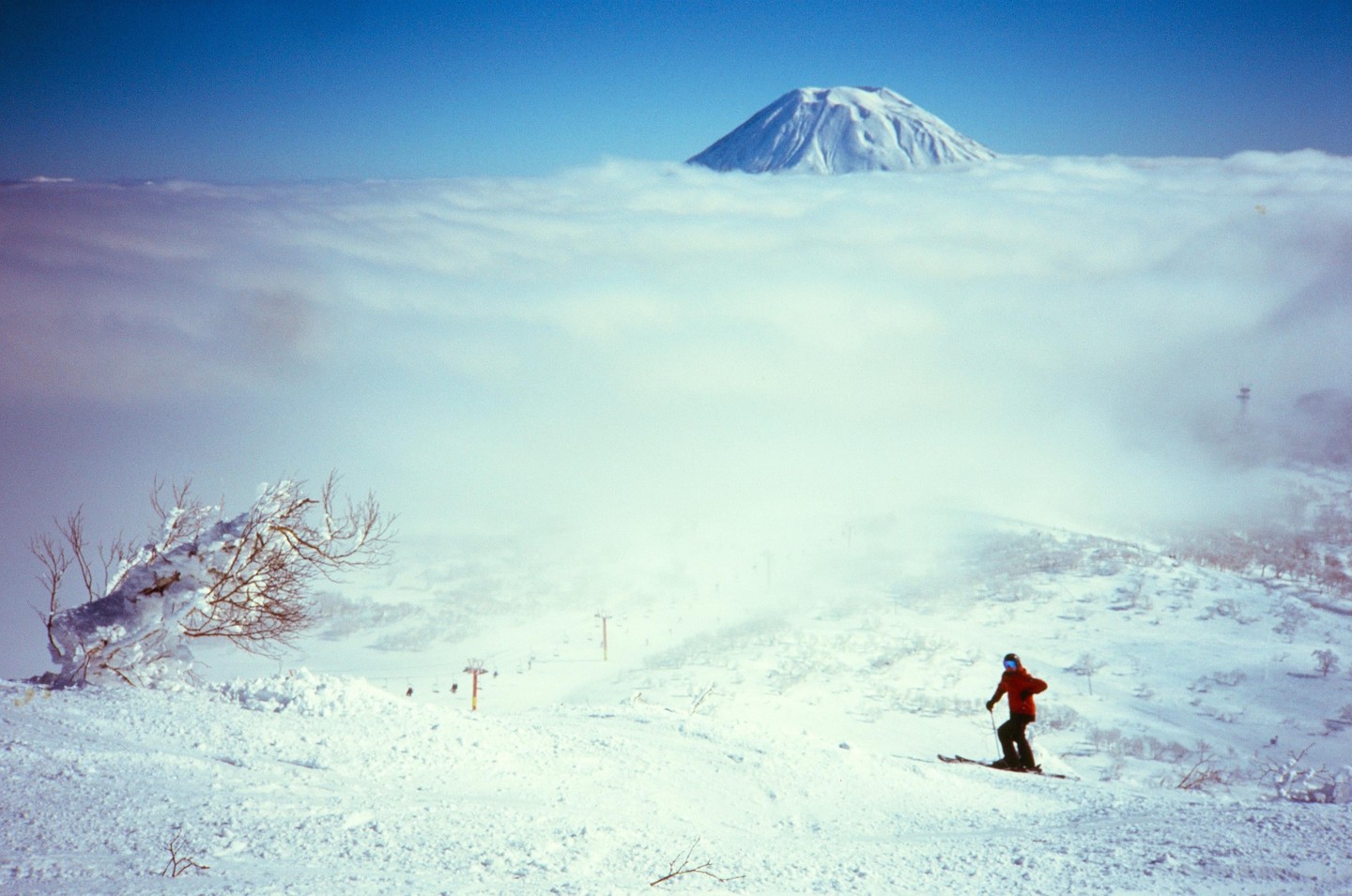 A skier descends a snowy mountain slope with a snow-covered mountain peak visible above a layer of clouds.