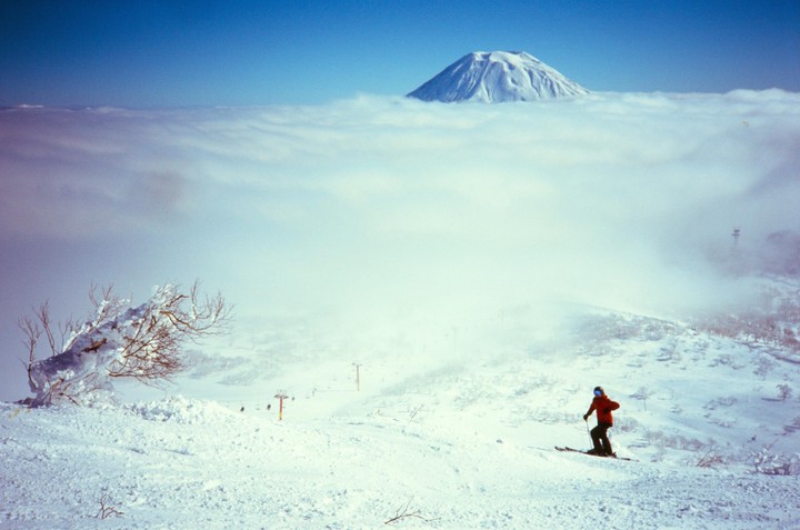 A skier descends a snowy mountain slope with a snow-covered mountain peak visible above a layer of clouds.