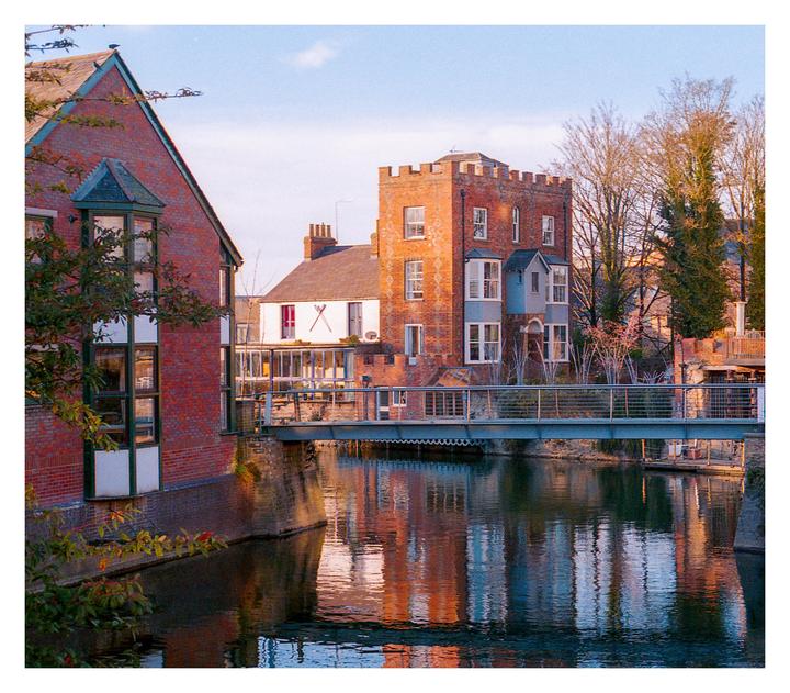 A brick building with a turret stands beside a river with a pedestrian bridge crossing it.