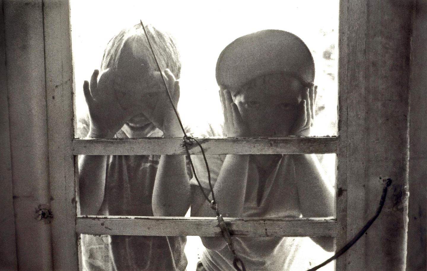 Two children are looking through a screen door, one with hands covering their face and the other with hands on their cheeks.