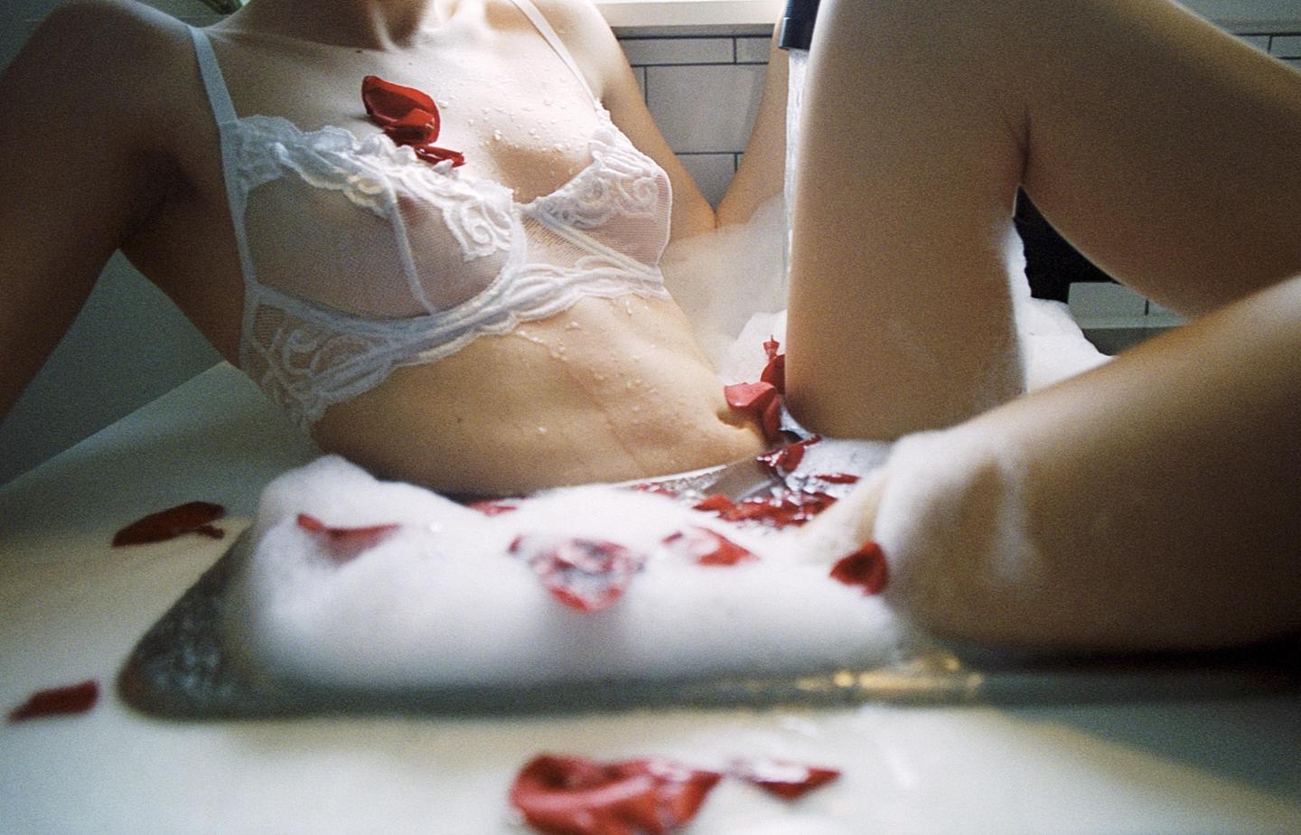 A woman in a white lace bra is in a bubble bath with red rose petals.