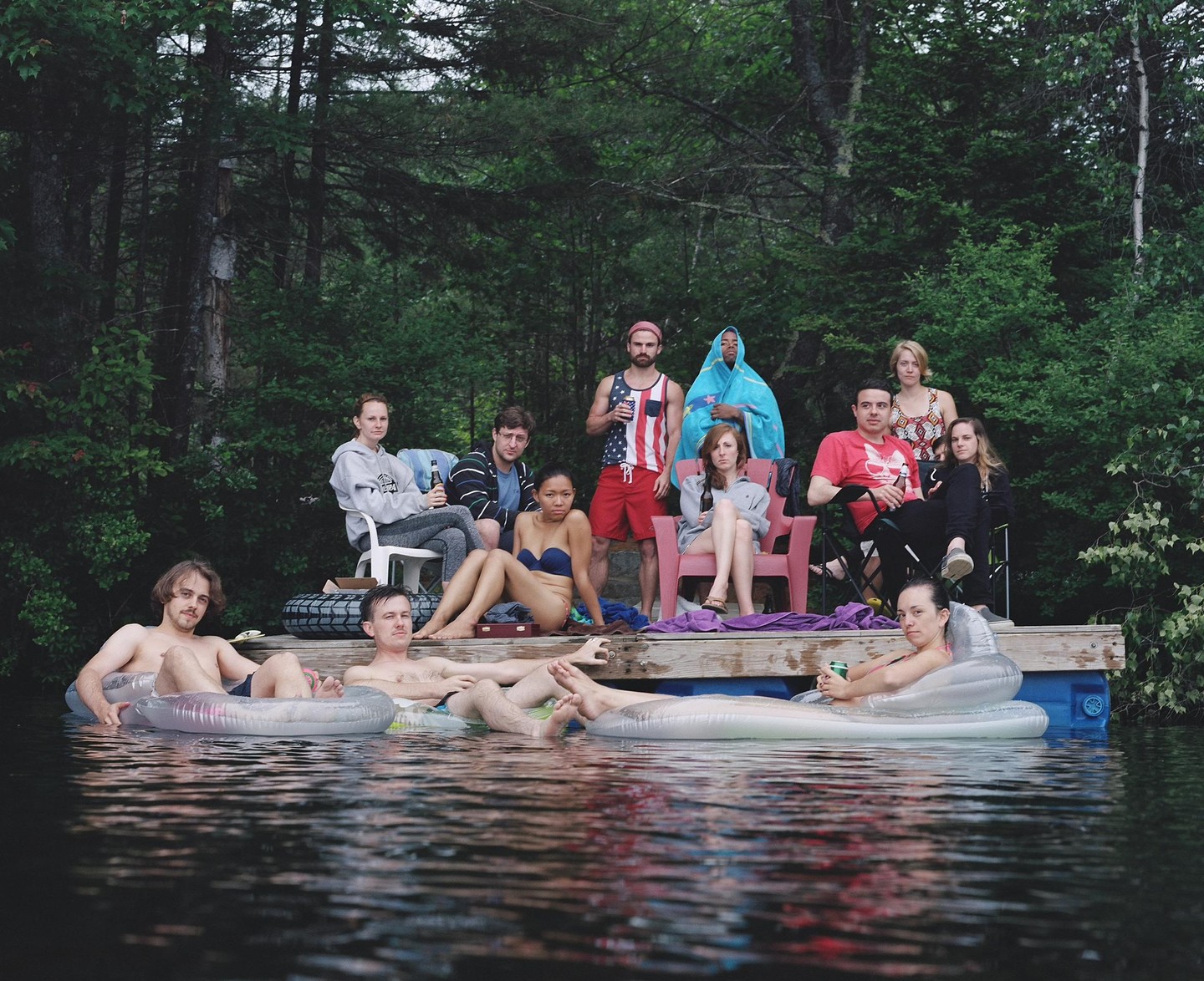 A group of friends relaxes on a dock and in the water in front of a dense forest.
