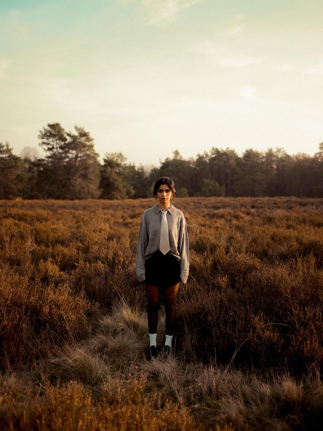 A woman stands in a field of tall, dry grass with trees in the background under a golden hour sky.
