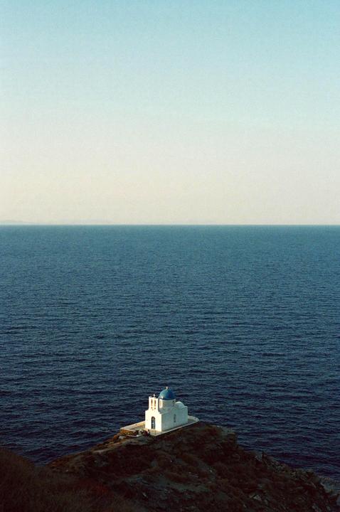 A white church with a blue dome sits on a cliff overlooking the ocean.
