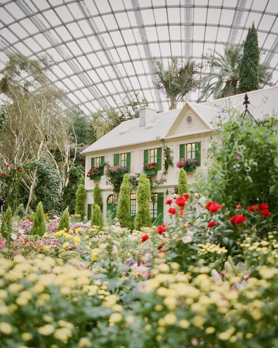 A house with green shutters and flower boxes is surrounded by a colorful garden under a glass dome.