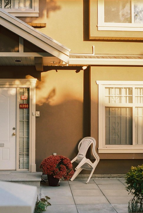 A white door and window are next to a potted plant and two stacked chairs outside a building.