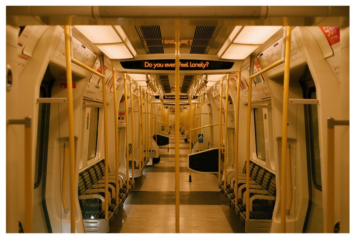 An empty subway car with yellow poles and seats is illuminated by overhead lights, with a digital sign displaying the question "Do you ever feel lonely?".