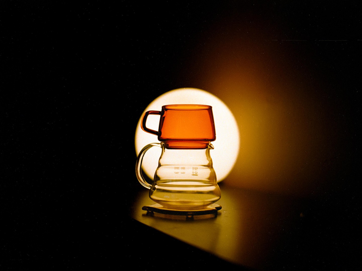 A glass coffee carafe with an orange glass cup on top is illuminated by a circular light source in the background.