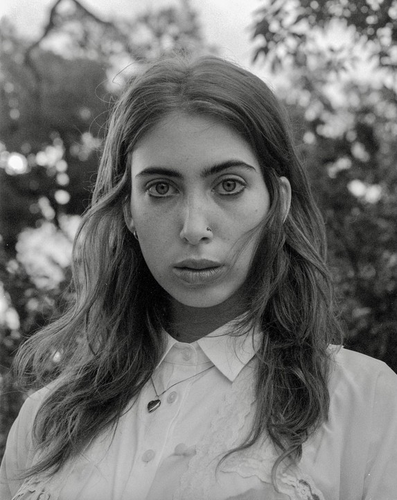 A woman with long hair and a nose piercing looks directly at the viewer.